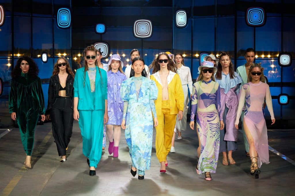 un groupe d’individus sur un podium de mode, vêtus de tenues colorées, dans un décor éclairé par des lumières circulaires bleues, suggérant un défilé de mode.