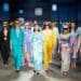 un groupe d’individus sur un podium de mode, vêtus de tenues colorées, dans un décor éclairé par des lumières circulaires bleues, suggérant un défilé de mode.